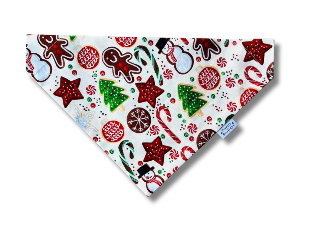 A triangular pet bandana with a festive Christmas print featuring candy canes, gingerbread men, stars, snowmen, and ornaments on a white background, with a small blue 'Peanut' brand tag.