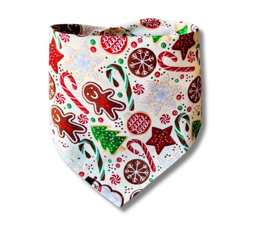 A triangular pet bandana featuring a festive Christmas print with gingerbread men, candy canes, snowflakes, stars, and trees on a cream background, shown on a white studio background with a soft drop shadow.
